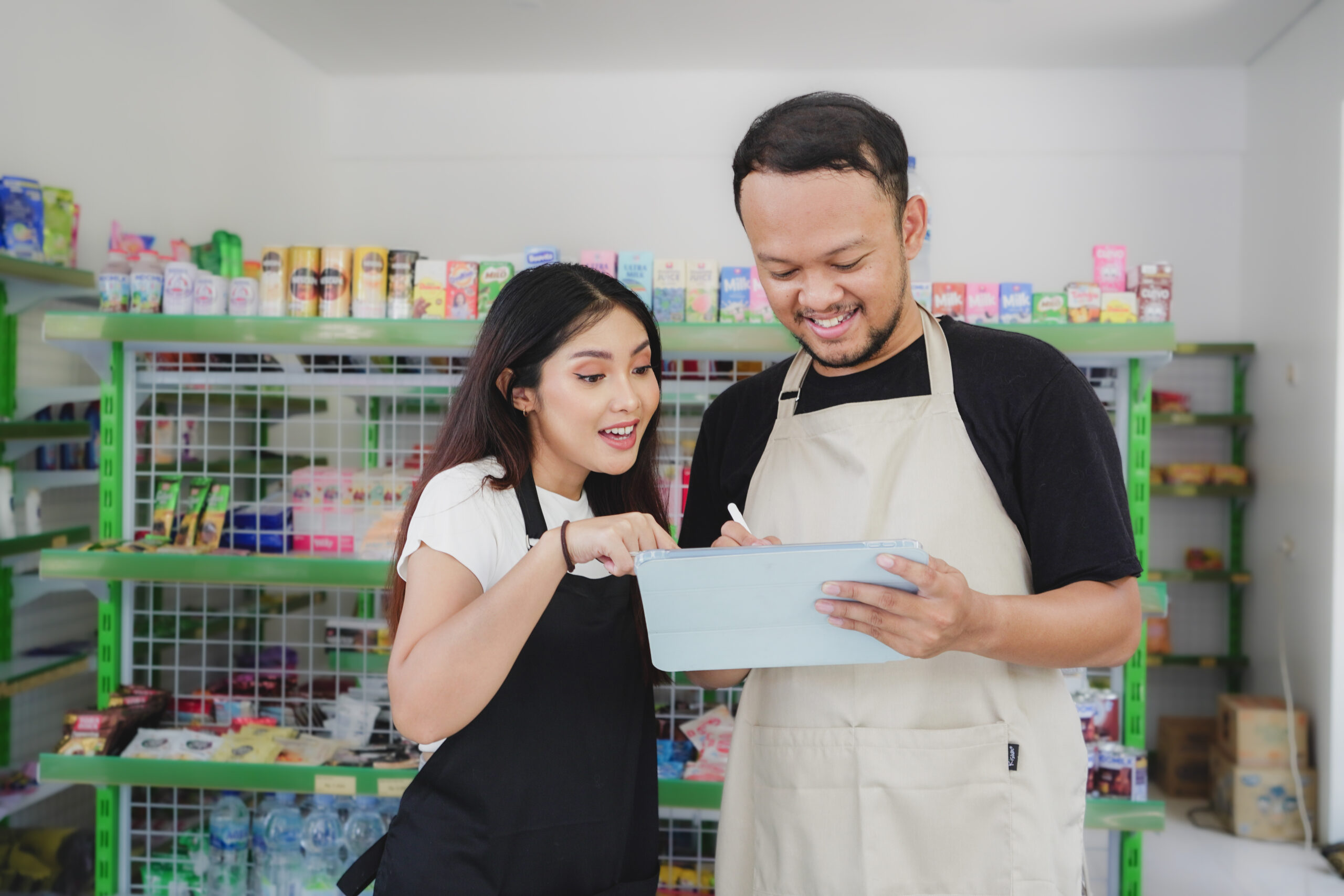Smiling Asian people holding a tablet, cashier is wearing black and cream apron standing in a groceries/convenient store